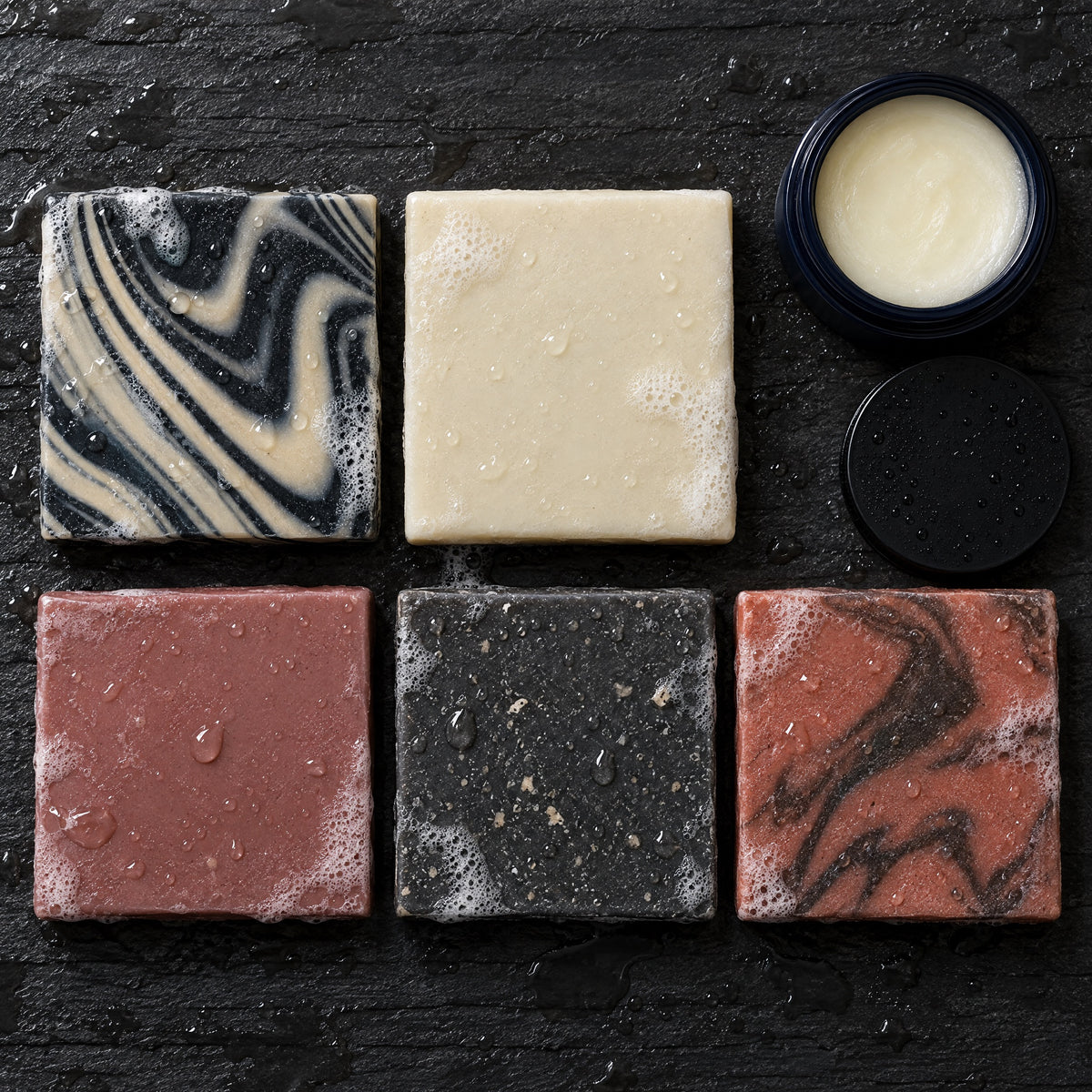 Overhead view of the five Old Guard soap bars and an open jar of Balm of the Brave on wet slate