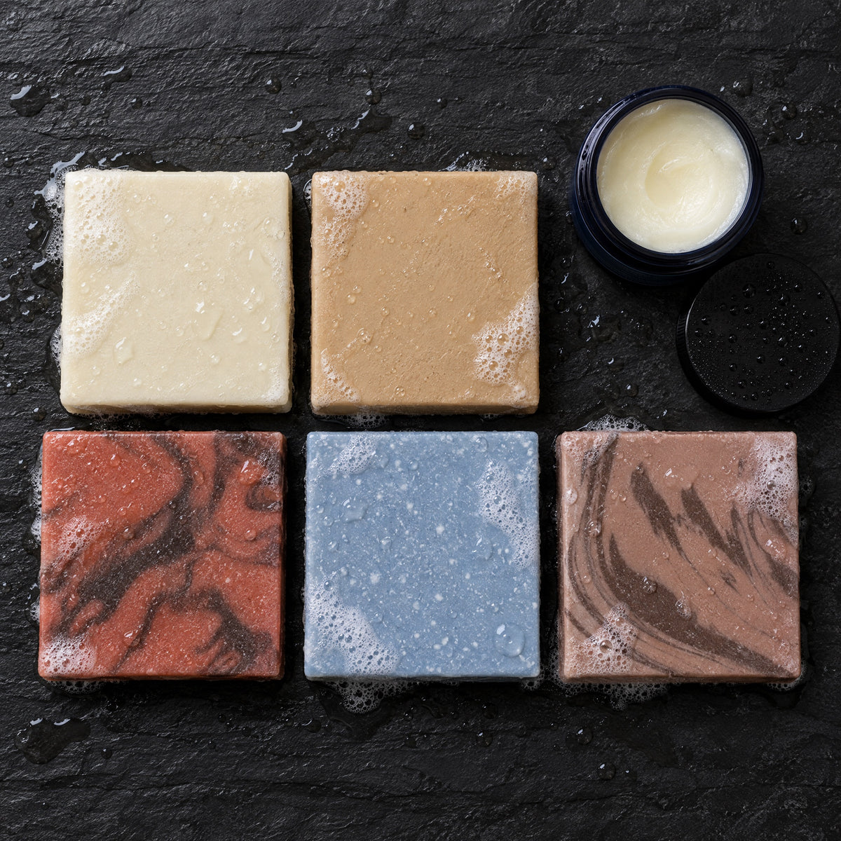 Overhead view of the five Tallow Reserve soap bars and an open jar of Balm of the Brave on wet slate