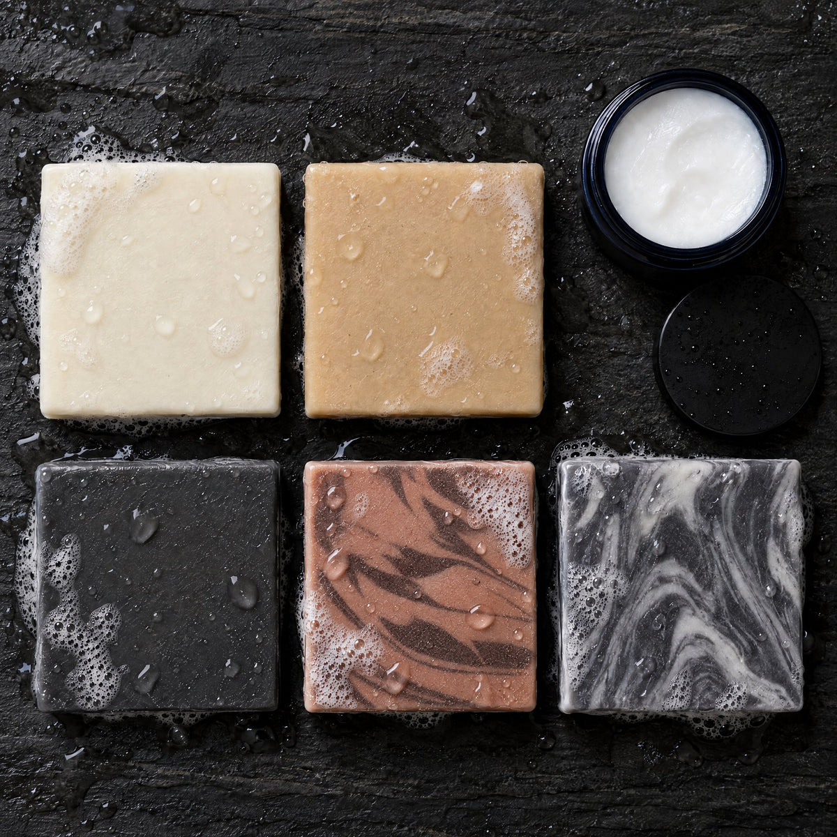 Overhead view of the five Timberline soap bars, including Sweet Sawmill Timber, and an open jar of Balm of the Brave on wet slate